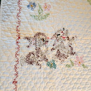 May include: Cream-colored, quilted textile with embroidered floral and geometric designs. The quilt features a repeating diamond pattern and a border with red stitching. The floral motifs include blue, pink, and green cross-stitch flowers.
