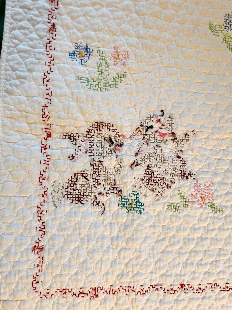 May include: Cream-colored, quilted textile with embroidered floral and geometric designs. The quilt features a repeating diamond pattern and a border with red stitching. The floral motifs include blue, pink, and green cross-stitch flowers.