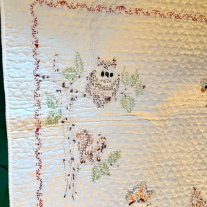 May include: Cream-colored quilt with embroidered owl and floral designs. The quilt features a red stitched border and a repeating diamond pattern. The owl is brown with black eyes, and the leaves are green.