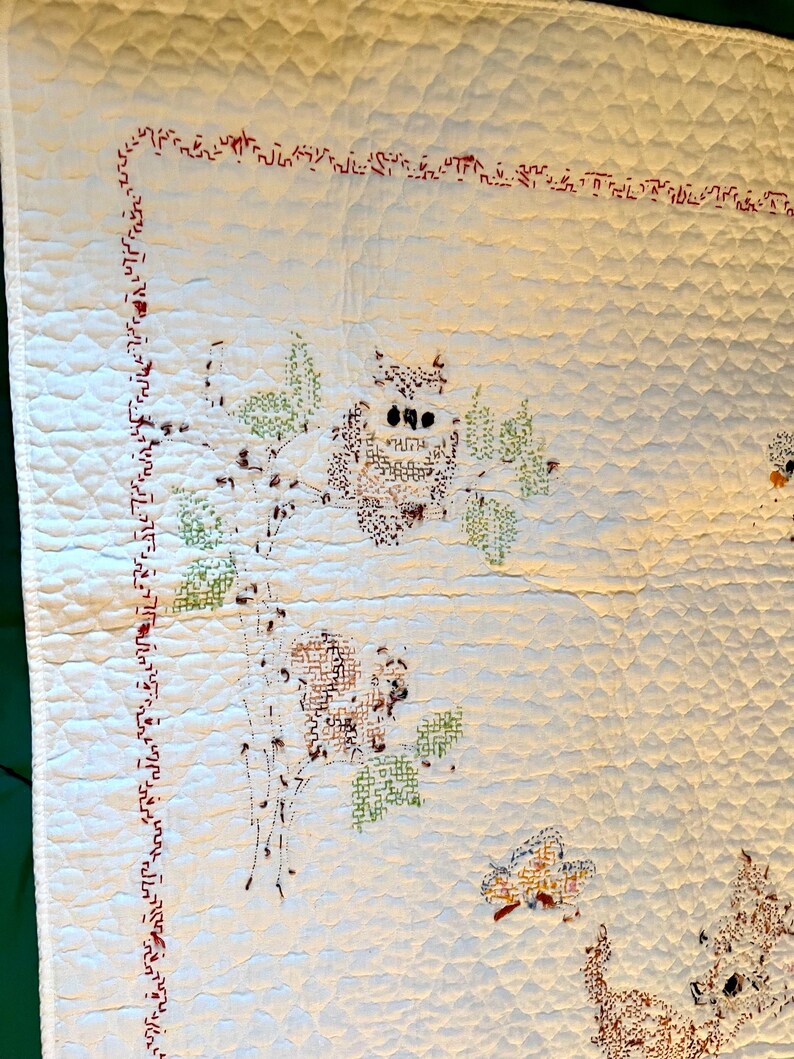 May include: Cream-colored quilt with embroidered owl and floral designs. The quilt features a red stitched border and a repeating diamond pattern. The owl is brown with black eyes, and the leaves are green.