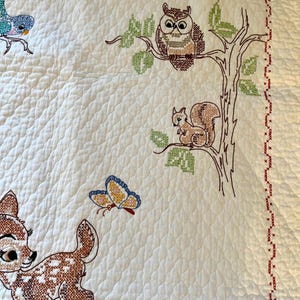 May include: White quilted blanket with embroidered woodland creatures. Features a deer, owl, squirrel, and butterfly in brown, blue, and yellow. The blanket has a red cross-stitch border and a floral pattern.