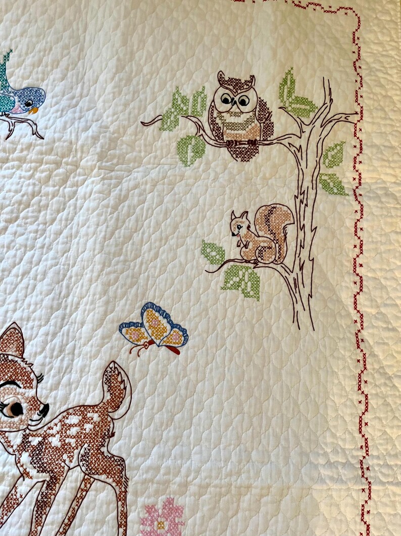 May include: White quilted blanket with embroidered woodland creatures. Features a deer, owl, squirrel, and butterfly in brown, blue, and yellow. The blanket has a red cross-stitch border and a floral pattern.