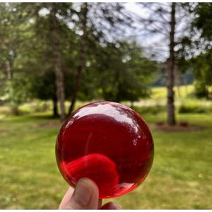 May include: A smooth, spherical object made of translucent red glass. The sphere has a polished, reflective surface that catches light, creating bright highlights across its rounded form. The deep red color is consistent throughout the glass.