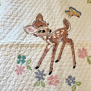 May include: Embroidered quilt featuring a fawn with brown and white spots, a butterfly, and cross-stitched flowers in pink, blue, and green. The quilt has a quilted pattern on a white background.