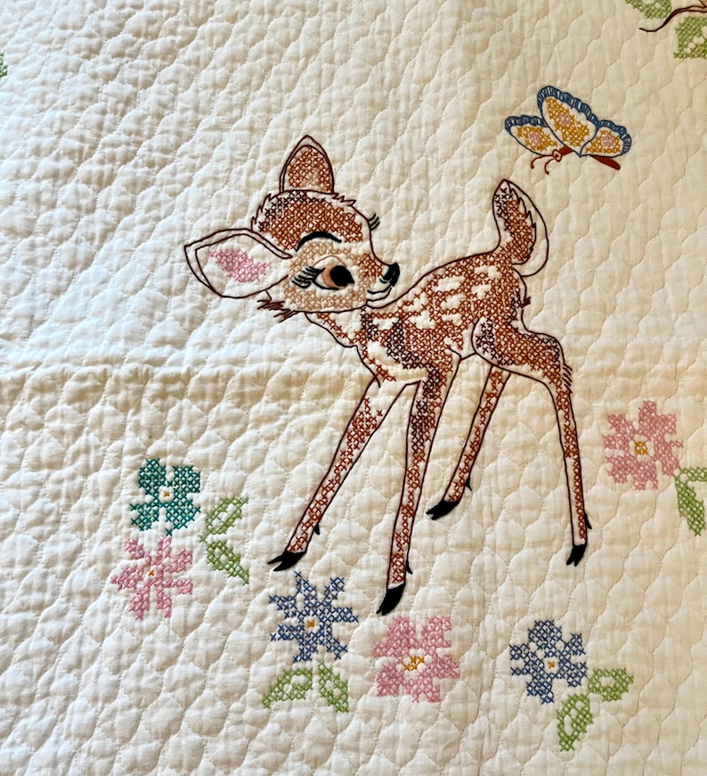 May include: Embroidered quilt featuring a fawn with brown and white spots, a butterfly, and cross-stitched flowers in pink, blue, and green. The quilt has a quilted pattern on a white background.