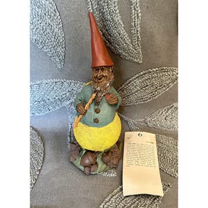 May include: A gnome figurine wearing a tall red hat, a teal jacket with brown buttons, and bright yellow trousers. The gnome holds a small candle and a heart, standing on a base with acorns and the name Mac inscribed on a rock, accompanied by a tag.
