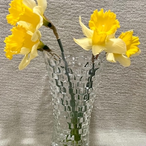 May include: A clear glass vase with a textured, square pattern holds several yellow daffodils. The flowers have bright yellow petals and green stems. The vase is approximately 8 inches tall.
