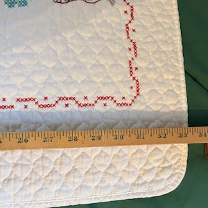 May include: White quilted fabric with red cross-stitch border and embroidered details. The design includes a brown hedgehog and turquoise floral elements. The fabric measures approximately 34 inches in length.