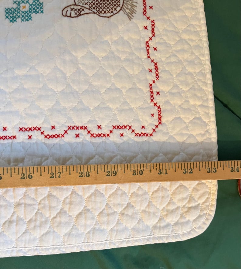May include: White quilted fabric with red cross-stitch border and embroidered details. The design includes a brown hedgehog and turquoise floral elements. The fabric measures approximately 34 inches in length.