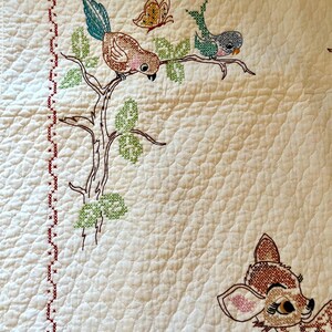 May include: Cream-colored quilt with embroidered woodland creatures. Features a brown bird with a teal tail, a butterfly, and a deer. The quilt has a red cross-stitch border and a quilted pattern.