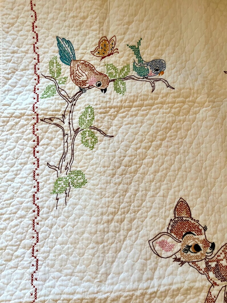 May include: Cream-colored quilt with embroidered woodland creatures. Features a brown bird with a teal tail, a butterfly, and a deer. The quilt has a red cross-stitch border and a quilted pattern.