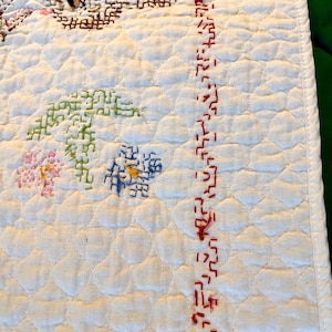 May include: Close-up of a white quilted textile with embroidered floral and geometric designs. The embroidery features pink, green, blue, and brown threads. A red stitched border runs along the right edge.