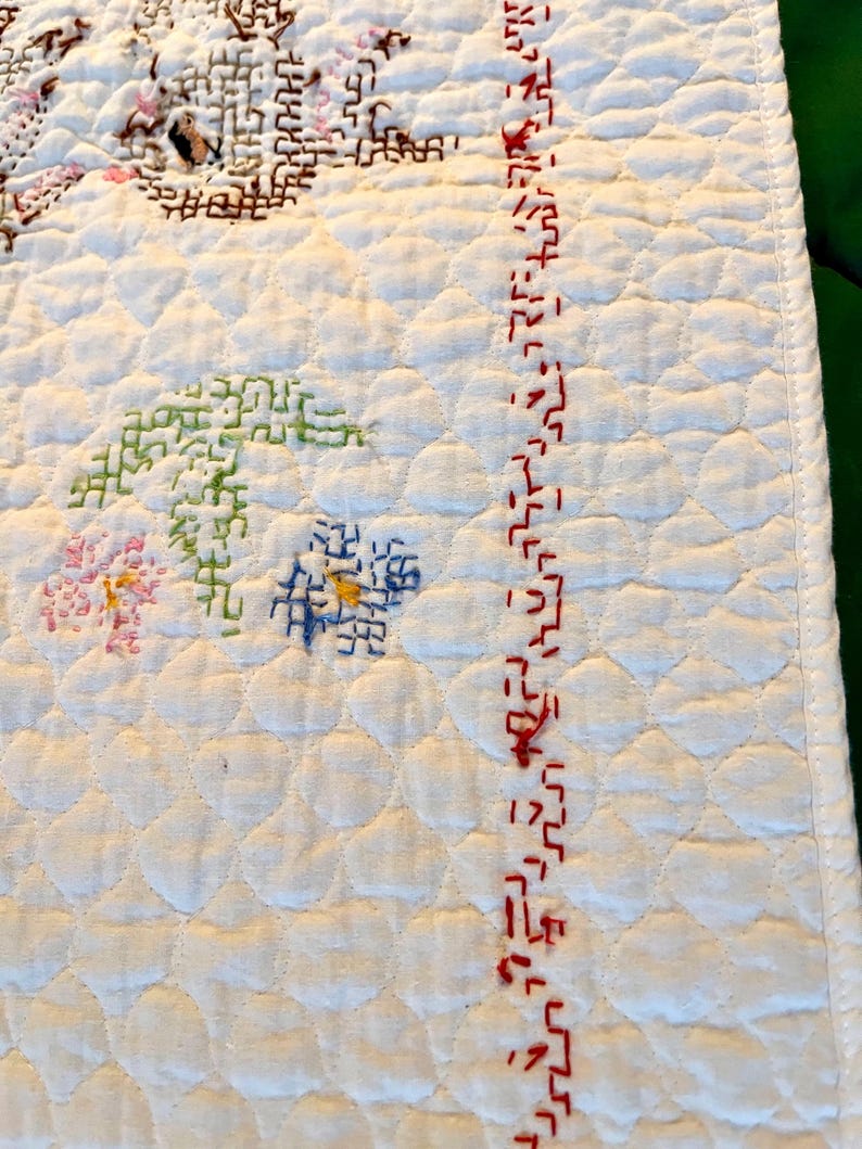 May include: Close-up of a white quilted textile with embroidered floral and geometric designs. The embroidery features pink, green, blue, and brown threads. A red stitched border runs along the right edge.