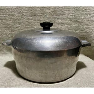 May include: A round, brushed aluminum cooking pot with a matching lid and a black knob handle. The pot features two side handles and a smooth, metallic finish with visible surface wear, showing a utilitarian design for stovetop cooking.