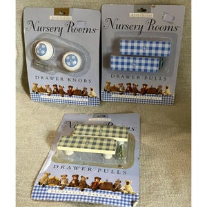 May include: Three packages of Anne Geddes Nursery Rooms decorative hardware. One package contains two round white drawer knobs with blue gingham centers. Two packages contain two rectangular drawer pulls each, one with blue and one with tan gingham.
