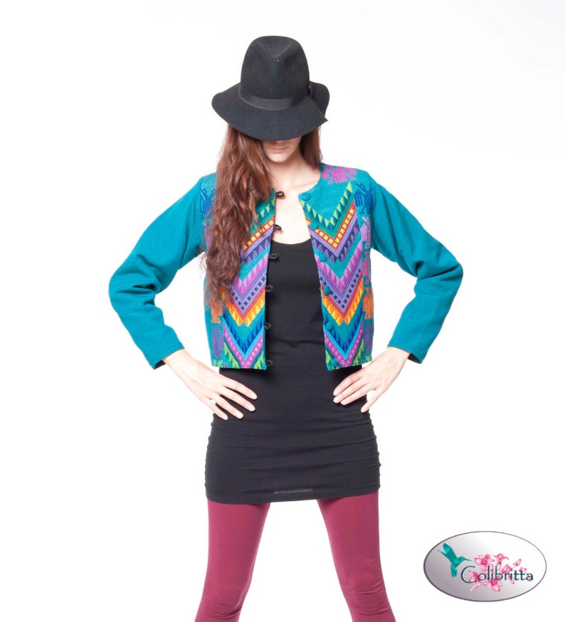Peut inclure: Une veste bleu turquoise avec un motif g&eacute;om&eacute;trique color&eacute; sur le devant. La veste a une fermeture &agrave; boutons et des manches longues. Le mannequin porte un d&eacute;bardeur noir, un legging bordeaux et un fedora noir.