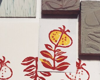 May include: Red and yellow hand-printed floral design on white paper. The design features a stylised flower with a red stem and leaves and a yellow centre. The print is surrounded by a white border.