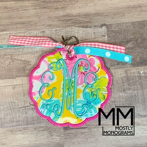 May include: A floral monogram keychain with a pink scalloped edge. The keychain features a light blue embroidered monogram on a colorful floral background. It is adorned with pink gingham and light blue polka dot ribbons.