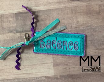 May include: A personalized teal and purple name tag with the name "Badence" embroidered in purple script. The tag has a teal background with a dotted border and is attached to a silver clip with teal and purple ribbons.
