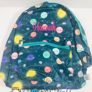 May include: A teal blue backpack with a colourful planets and stars print. The backpack has a front pocket and a zipper closure. The name "Hannah" is embroidered on the front of the backpack in pink.