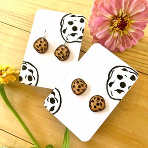 May include: Wooden cookie-shaped earrings and studs displayed on white cards with black and white abstract designs. The earrings and studs are brown with dark brown spots. A pink and yellow flower are in the background.