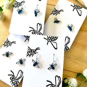 May include: Three sets of earrings featuring small, blue, translucent bee-shaped charms. Each pair is displayed on a white card printed with black line-art illustrations of bees. The earrings include silver-toned metal hooks or posts.