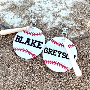 May include: Two white baseball-themed keychains with black stitching and red laces. One keychain has the name "BLAKE" and the other has the name "GREYSOU". Each keychain has a small wooden baseball bat attached.
