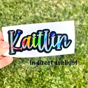 May include: A custom name sticker featuring the name Kaitlin in a bold, cursive font. The letters have a holographic, multi-colored finish that shifts in the light, outlined by a thick black border on a white rectangular backing sheet.