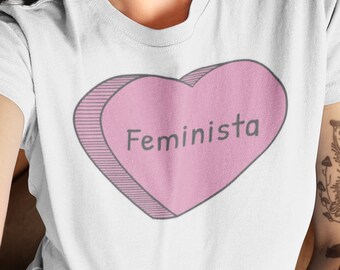 May include: White t-shirt with a pink heart graphic that says "Feminista" in black text.