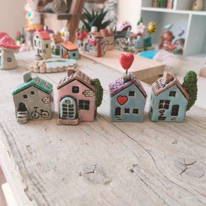 Könnte beinhalten: Eine Sammlung von Miniatur-Keramikhäusern in verschiedenen Farben, darunter Grau, Rosa und Blau, mit dekorativen Details wie einem Fahrrad, einem Herz und Bäumen. Die Häuser sind auf einer Holzoberfläche angeordnet, mit weiteren ähnlichen Häusern im Hintergrund.