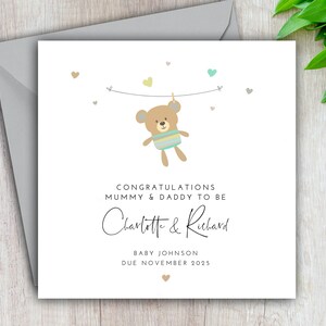 May include: A white card with a cute brown teddy bear hanging from a string with colorful hearts. The card says "Congratulations Mummy & Daddy to be Charlotte & Richard Baby Johnson Due November 2025".