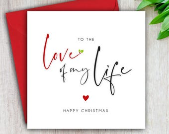Op de afbeelding: Een witte kerstkaart met de tekst "To the love of my life" in rode en zwarte letters, een klein rood hart en de woorden "Happy Christmas" eronder. De kaart zit in een rode envelop.
