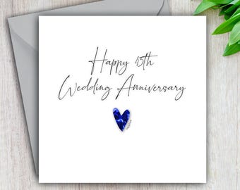 May include: A white greeting card with the text "Happy 45th Wedding Anniversary" in black script. A blue heart-shaped design is below the text. The card is inside a gray envelope.