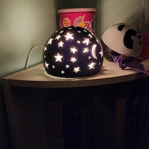 May include: A black nightlight shaped like a sphere with cutouts of stars and a crescent moon. The light is on and illuminating the stars. The nightlight is sitting on a white shelf with books below.