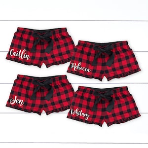 May include: Four pairs of red and black plaid pajama shorts with black bows and ruffles. Each pair has a different name in white script: Caitlin, Rebecca, Jen, and Whitney. The shorts are displayed on a white wooden surface.