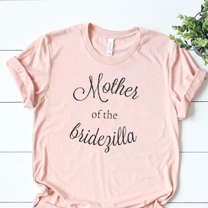 Puede incluir: Una camiseta rosa claro con el texto "Mother of the Bridezilla" en letras cursivas negras.