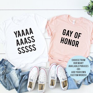 May include: Two t-shirts, one white and one pink, with text printed on them. The white t-shirt says "YAAAA AAASS SSSSS" and the pink t-shirt says "GAY OF HONOR". The text is in black. There are two pairs of white sneakers and a pair of blue jeans in the image.