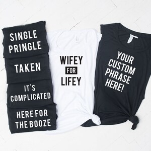May include: Five black tank tops with white text, reading "Single Pringle", "Taken", "It's Complicated", "Here for the Booze", and "Your Custom Phrase Here!"  A white tank top with black text reads "Wifey for Lifey".