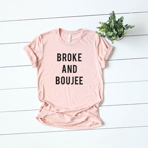 以下が含まれることがあります： 黒文字で「BROKE AND BOUJEE」とプリントされた薄いピンクのTシャツです。