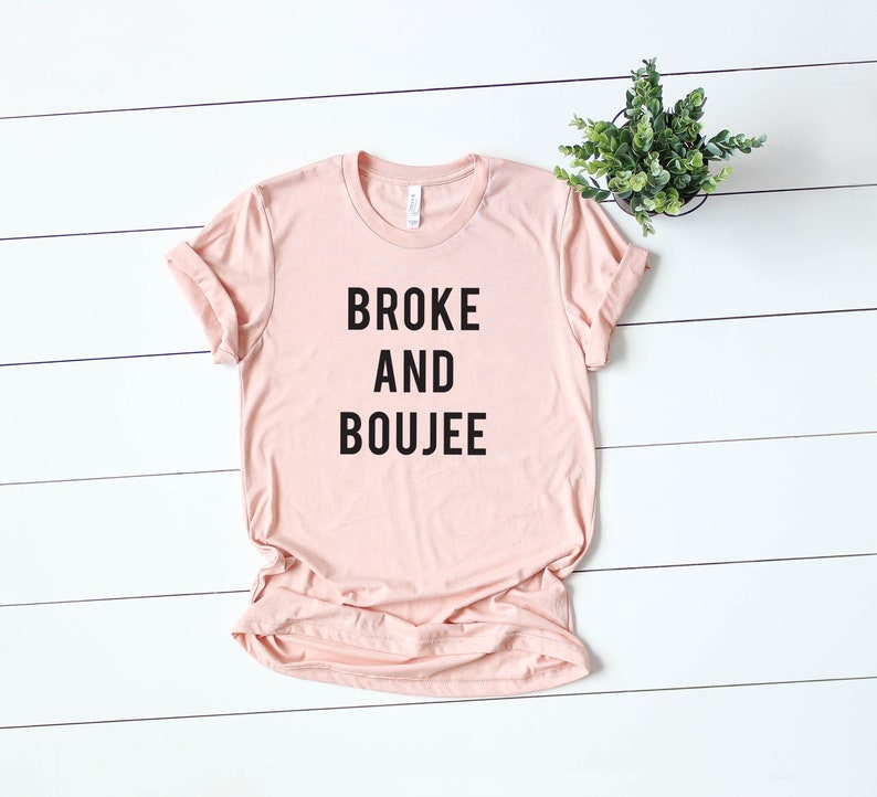 以下が含まれることがあります： 黒文字で「BROKE AND BOUJEE」とプリントされた薄いピンクのTシャツです。
