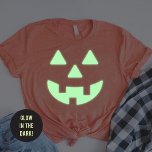 May include: A coral colored t-shirt with a glow-in-the-dark jack-o'-lantern face design. The text "Glow in the Dark!" is on a black circle.