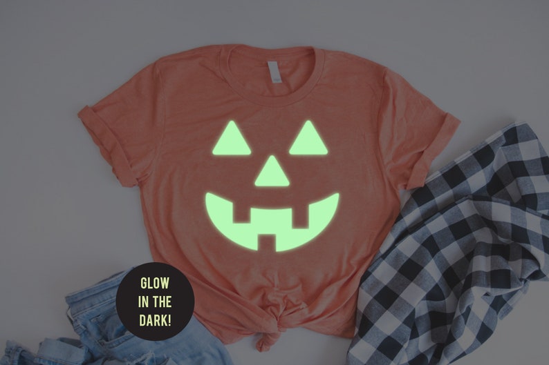 May include: A coral colored t-shirt with a glow-in-the-dark jack-o'-lantern face design. The text "Glow in the Dark!" is on a black circle.