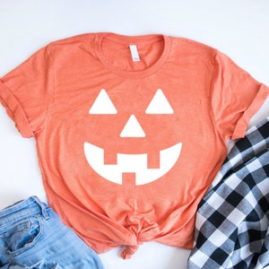 May include: A bright orange t-shirt with a white jack-o'-lantern face graphic. The shirt is tied in a knot at the bottom and is laying on a white surface with blue jeans and a black and white checkered fabric.