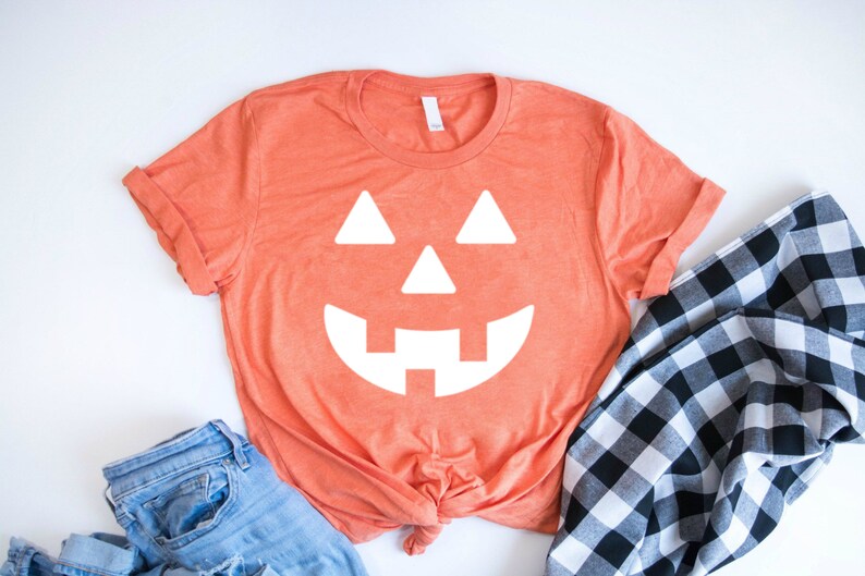 May include: A bright orange t-shirt with a white jack-o'-lantern face graphic. The shirt is tied in a knot at the bottom and is laying on a white surface with blue jeans and a black and white checkered fabric.