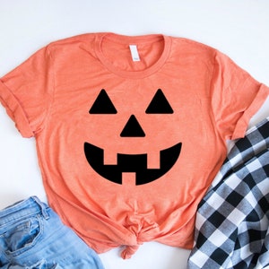 May include: A coral orange t-shirt with a black jack-o'-lantern face graphic. The shirt is made of a soft, comfortable material and is perfect for Halloween.
