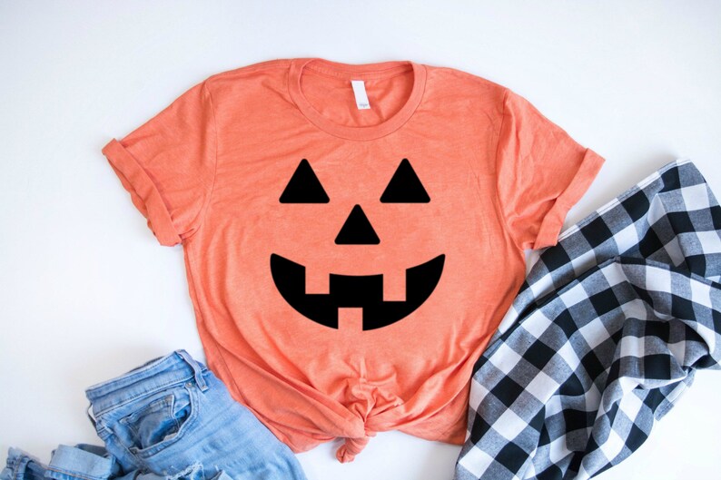 May include: A coral orange t-shirt with a black jack-o'-lantern face graphic. The shirt is made of a soft, comfortable material and is perfect for Halloween.