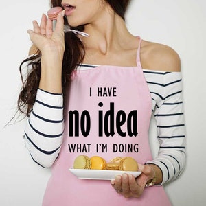 Puede incluir: Un delantal rosa con el texto "I HAVE no idea WHAT I'M DOING" se usa sobre una camisa de manga larga a rayas. Una persona sostiene un plato de macarons y se come uno. Los macarons son de varios colores.