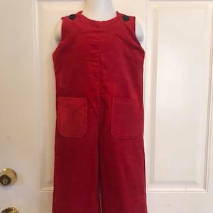 May include: A red corduroy overall with two front pockets and black buttons on the shoulders. The garment is sleeveless and hangs on a white mannequin. The overall is a solid, vibrant red.