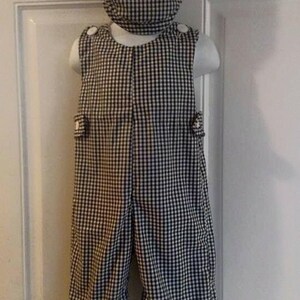 May include: A black and white gingham-patterned outfit for a child, including overalls and a matching cap. The overalls have button details on the shoulders and sides. The outfit is displayed on a white mannequin.
