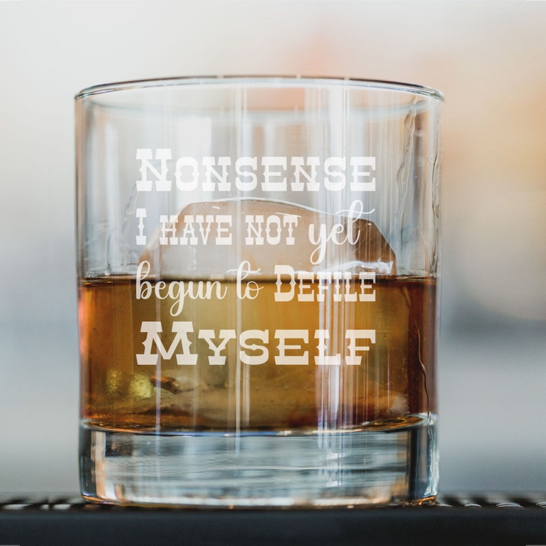 May include: A clear glass tumbler with a quote etched on the side: "Nonsense I have not yet begun to defile myself". The tumbler is filled with amber liquid and ice cubes.